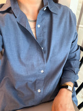Person wearing a blue shirt with visible buttons and a watch on a chair.